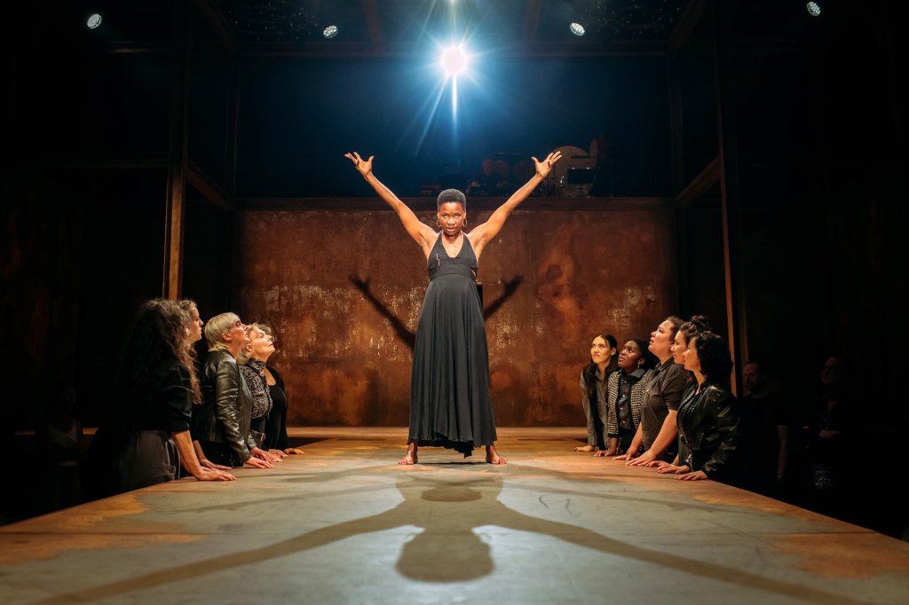 An actress stands in the middle of a runway holding her arms out overhead with a stern expression on her face, 10 women stand below to either side with their hands on the runway, looking up at the actress