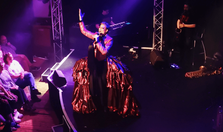 Reuben Kaye stands on a stage with a big shiny red bustle on his hips, singing while holding a hand up in the air