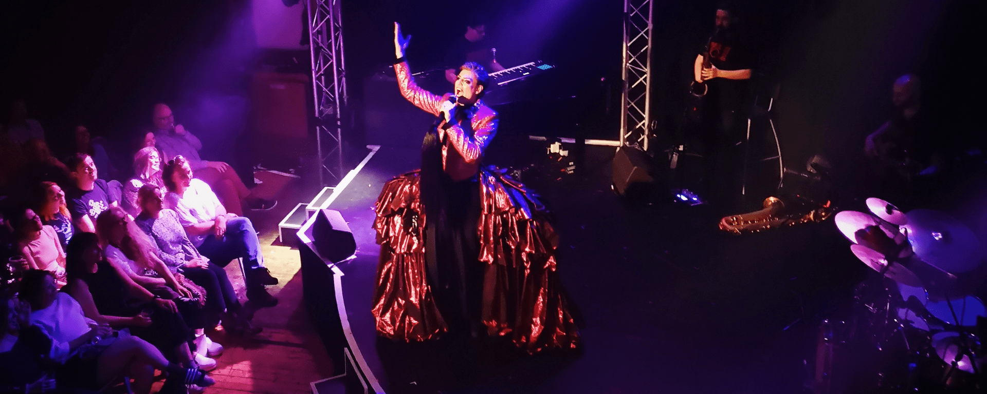 Reuben Kaye stands on a stage with a big shiny red bustle on his hips, singing while holding a hand up in the air