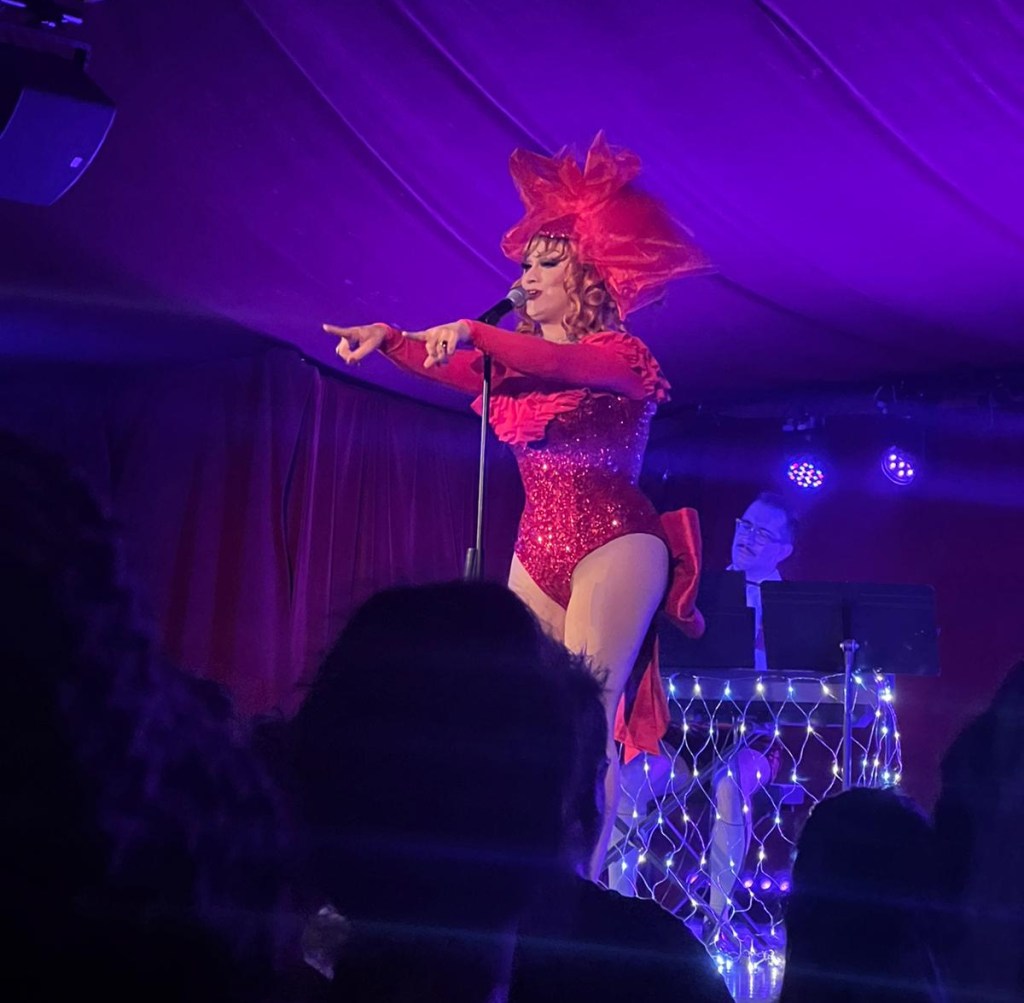 Jinkx sings into a microphone while pointing both fingers out in front of her toward the crowd