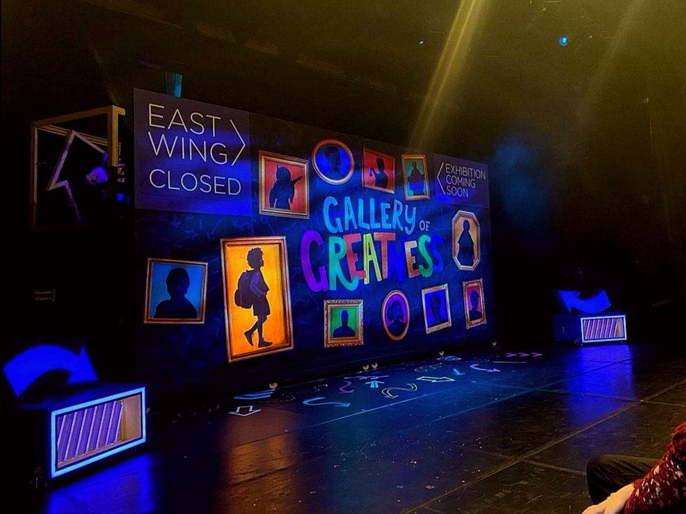 A picture of the stage, on which there is a wall with lots of frames showing silhouettes of women from history. There is writing that says Gallery of Greatness