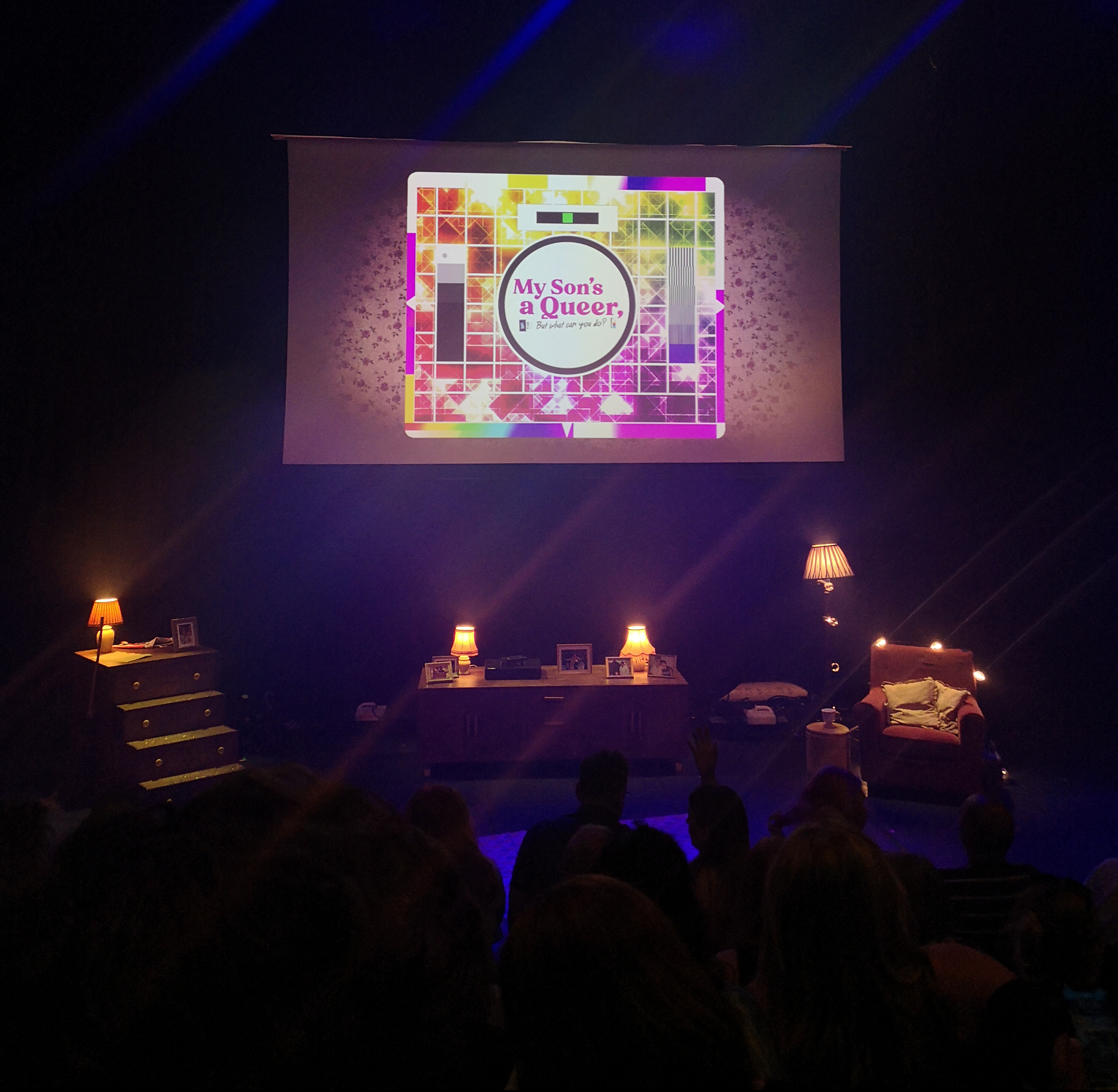 On a projected screen are the words My Son's a Queer but what can you do? and below there is a minimal set of a living room with an armchair, lamps and a cabinet