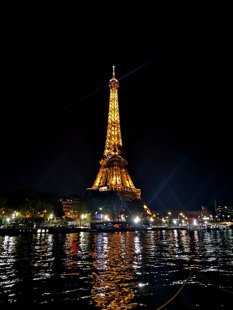 The Eiffel tower is light up and glowing in the night sky, its lights reflected in the Seine river