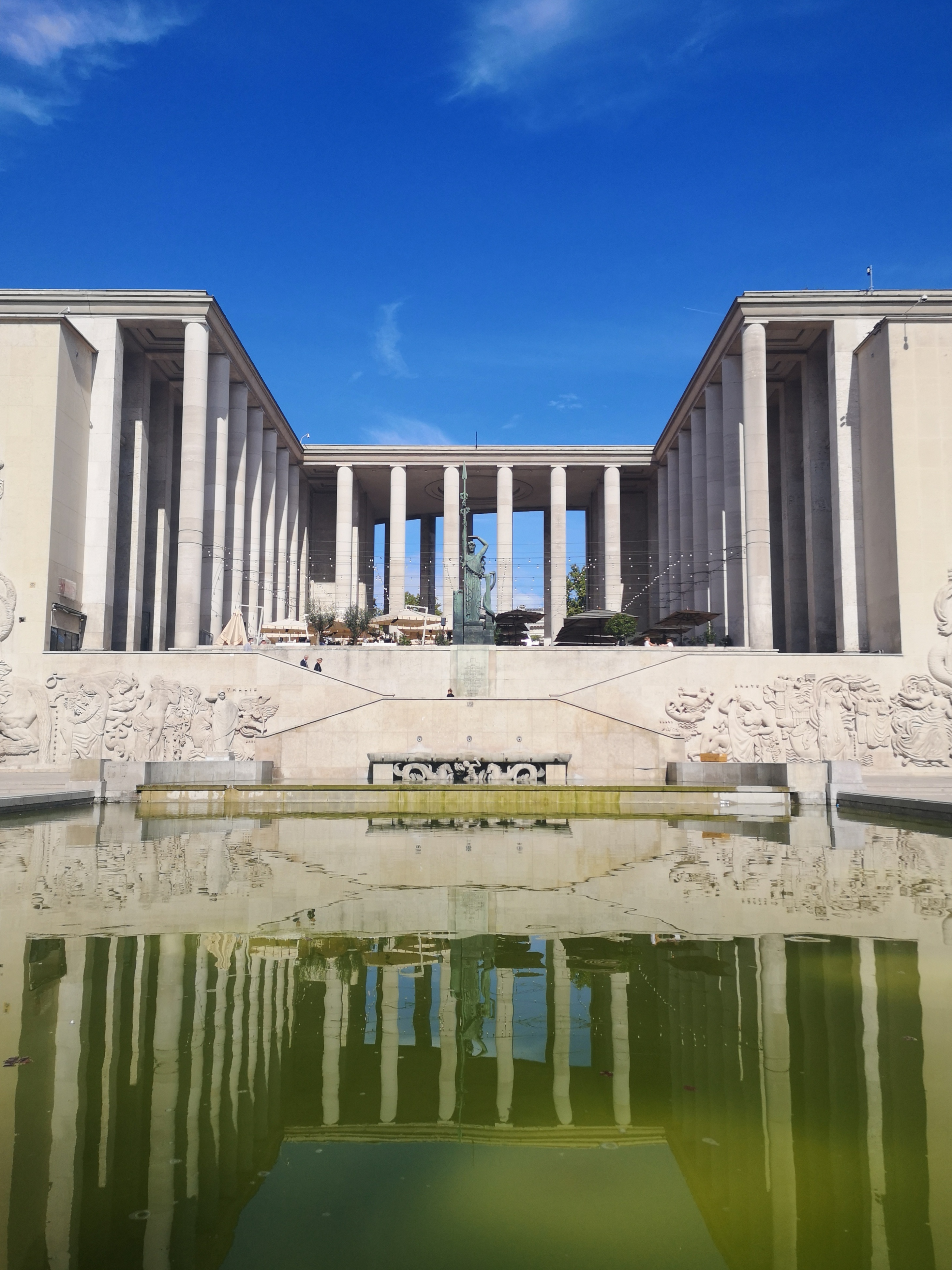 A greek looking building with columns shines bright in the sunshine, reflecting on a green pond that stretches out in front of it and the sky is a gorgeous blue