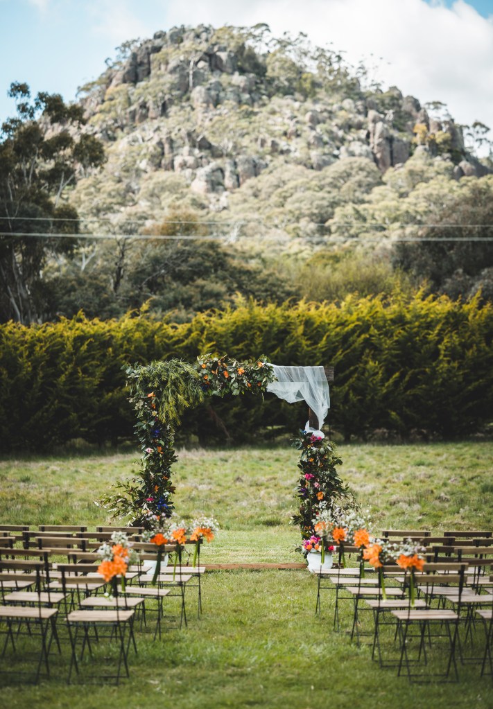 The wedding set up, a leafy flowery arch stands in front of rows of chairs, in the background is a massive rocky mountain decorated with trees