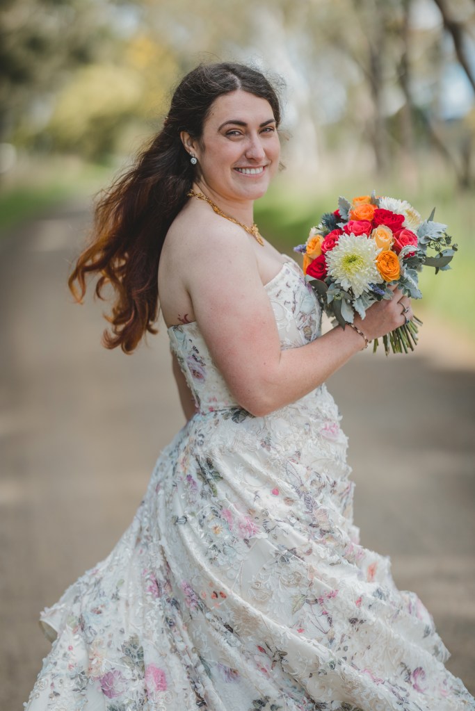 Georgia wears a white dress with green and pink flower details that moves while she spins around, she is smiling and holding a bouquet of white and orange flowers