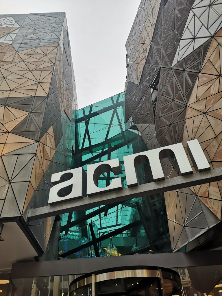 In a patterned brown metal and glass building there is a sign that reads 'acmi'