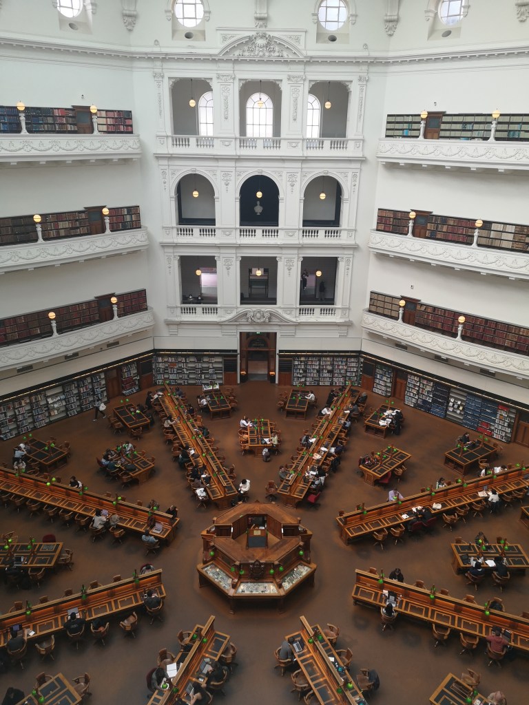 A stunning library space, rows of desks fan out from a circular desk in the centre of the room, three floors of windows and shelves tower above toward a domed ceiling
