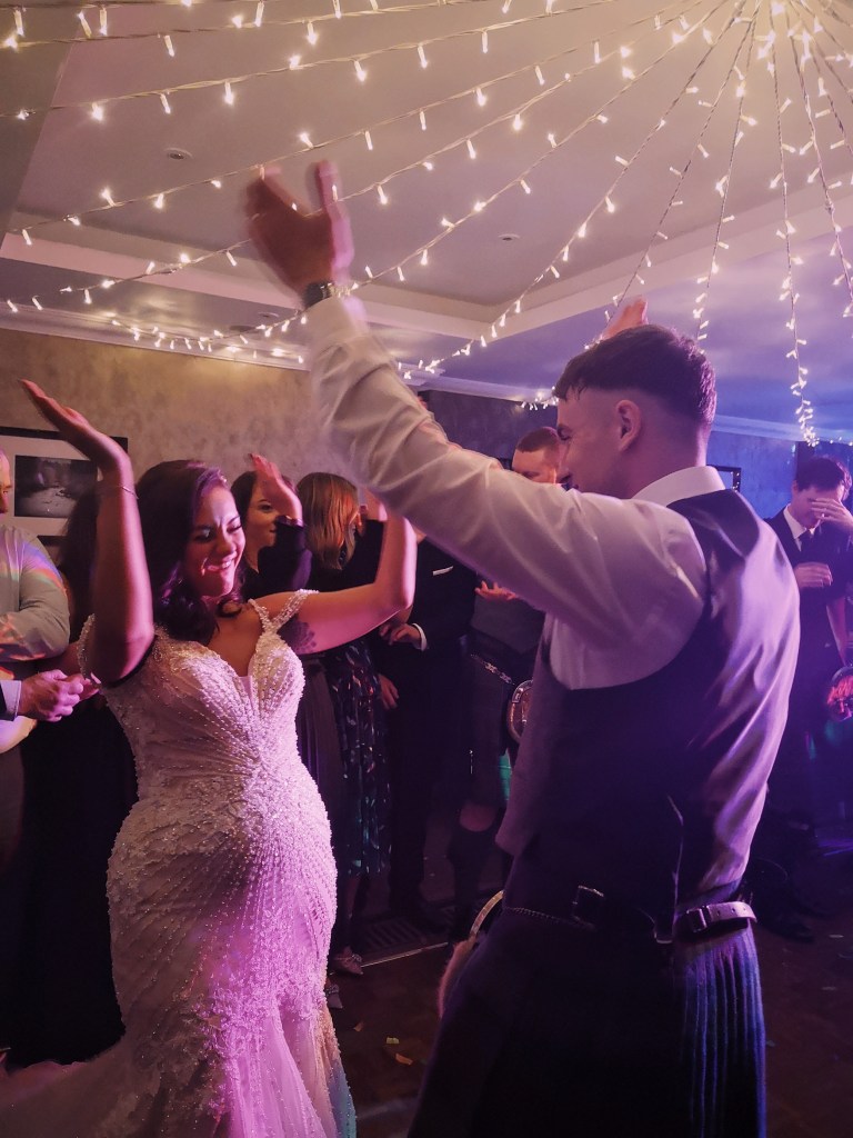 A bridge and groom on the dancefloor smiling at each other as they hold their hands up to clap overhead