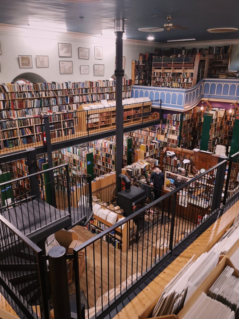 A beautiful bookshop with two floors stacked to the ceiling with books, there is a log burner in the middle of the room