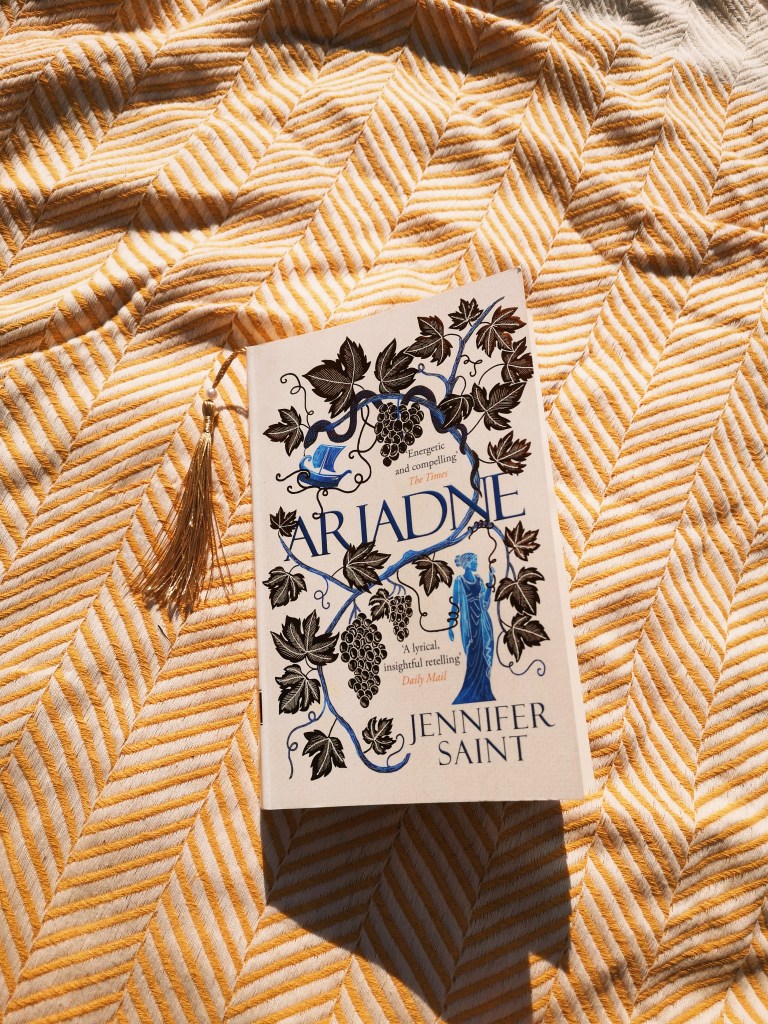 A copy of Ariadne by Jennifer Saint shines in the sun on a picnic blanket