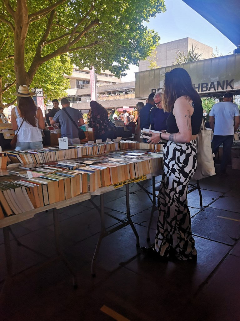A girl in funky trousers stands in front of a table stacked with books, browsing