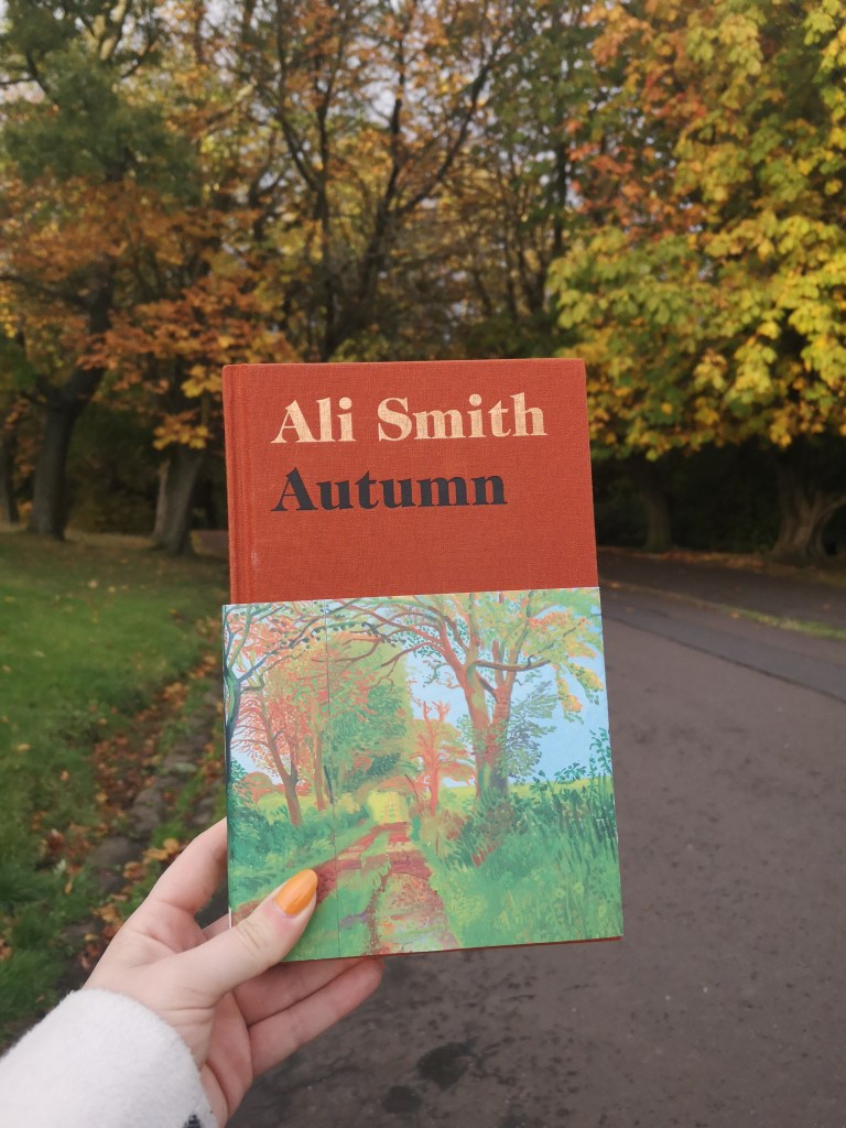 A copy of Ali Smith's autumn held up in front of real autumn trees