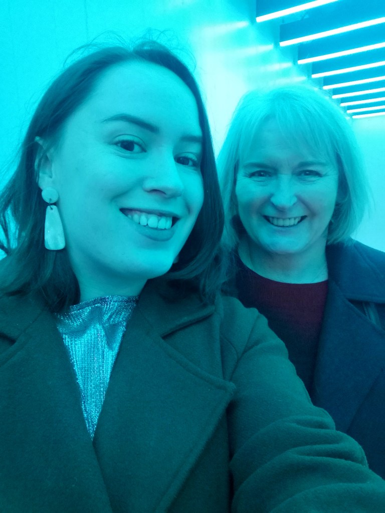 Two people, a young woman and her middle aged mother smile for a selfie, illuminated in pale blue light