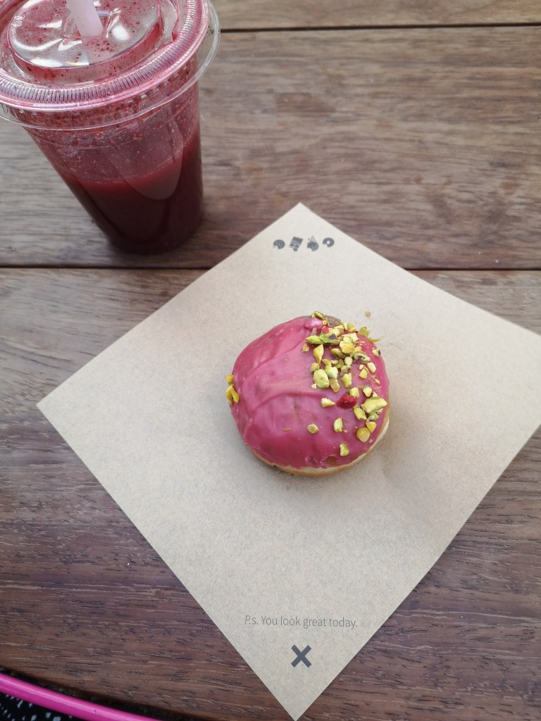 a mini pink donut sits on a piece of brown paper beside a berry smoothie in a cup