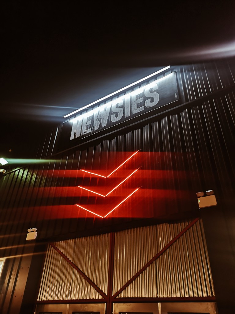 A sign saying Newsies is lit up and beneath it is three red neon arrows pointing down at the door