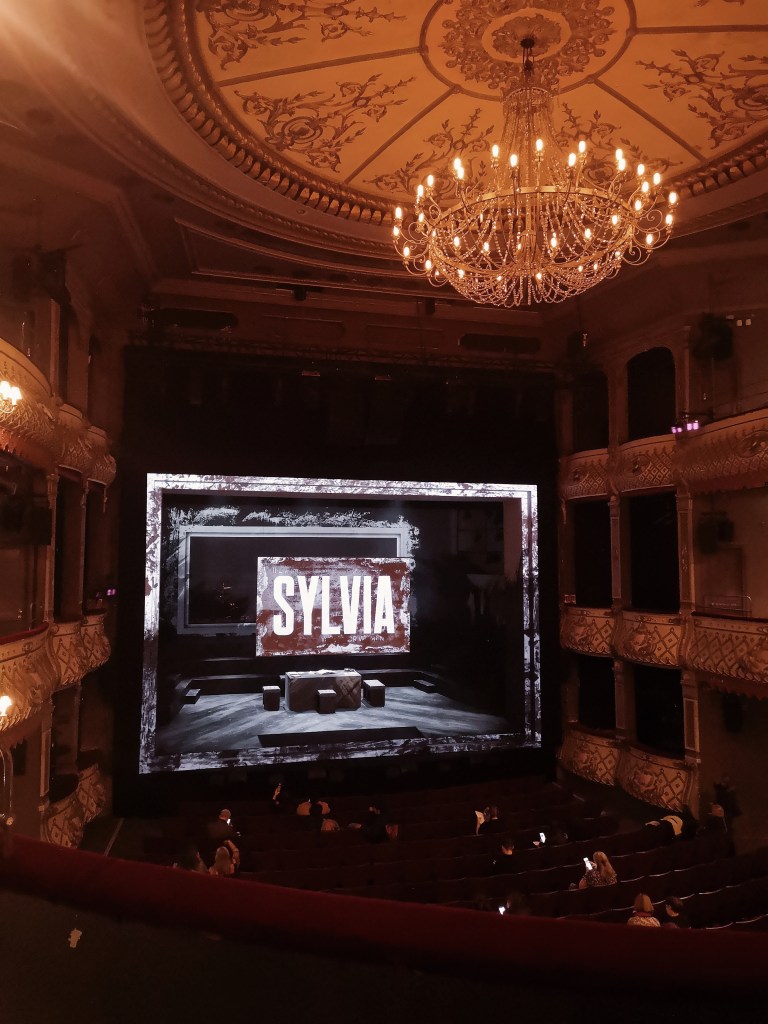 Interior of the Old Vic theatre with a chandelier and the word Sylvia projected on stage