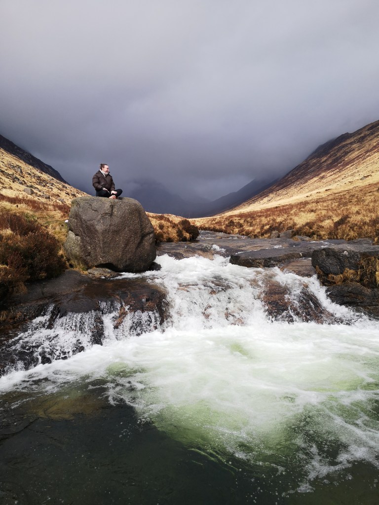 In a valley Helen sits on top of a giant boulder beside a rushing stream of water
