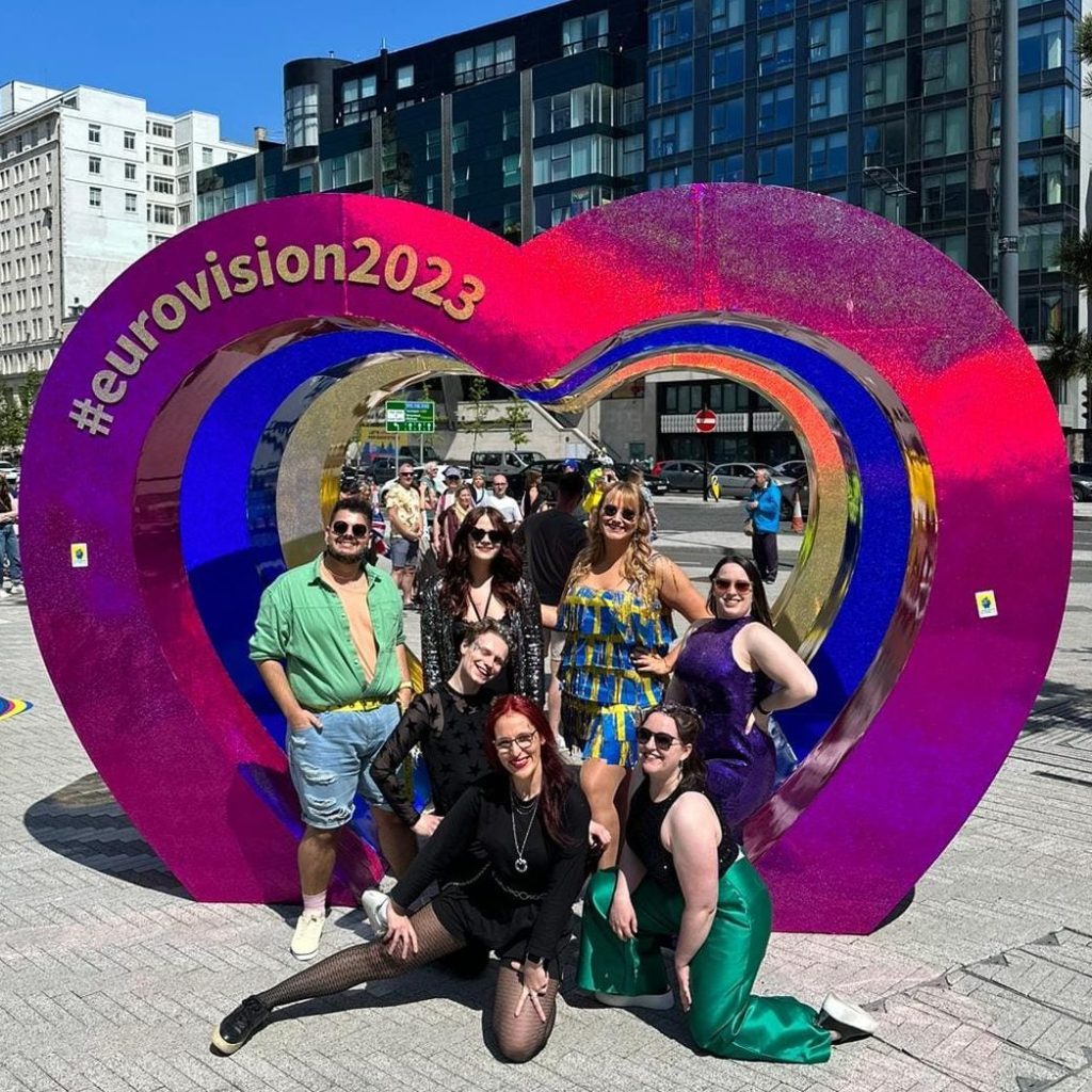 Surrounded by the outline of a giant pink heart, seven total babes pose together, smiling