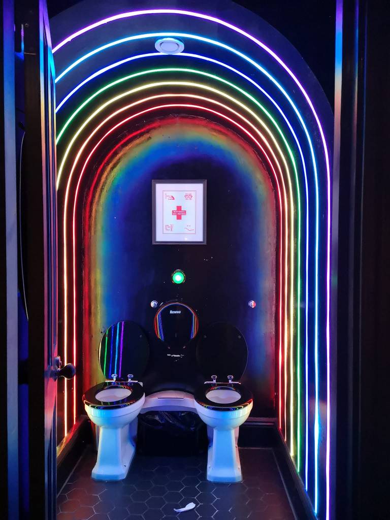Two toilets sit side by side in one stall, framed by rainbow neon lights