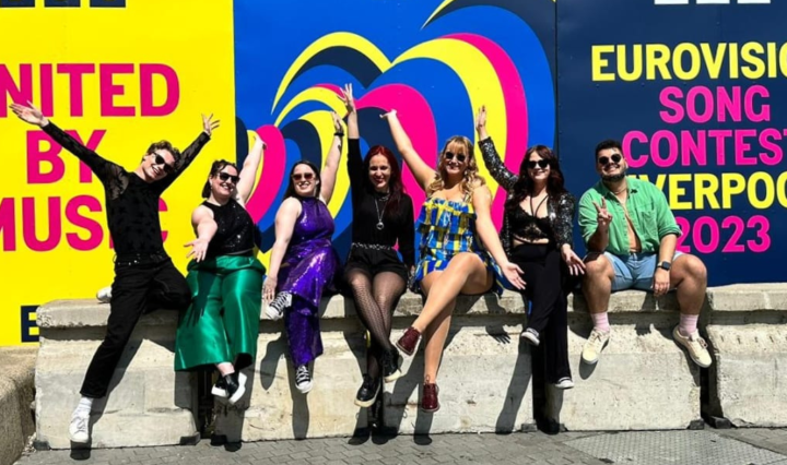 Seven lovely people sit on a wall and smile with their arms stretched out fabulously