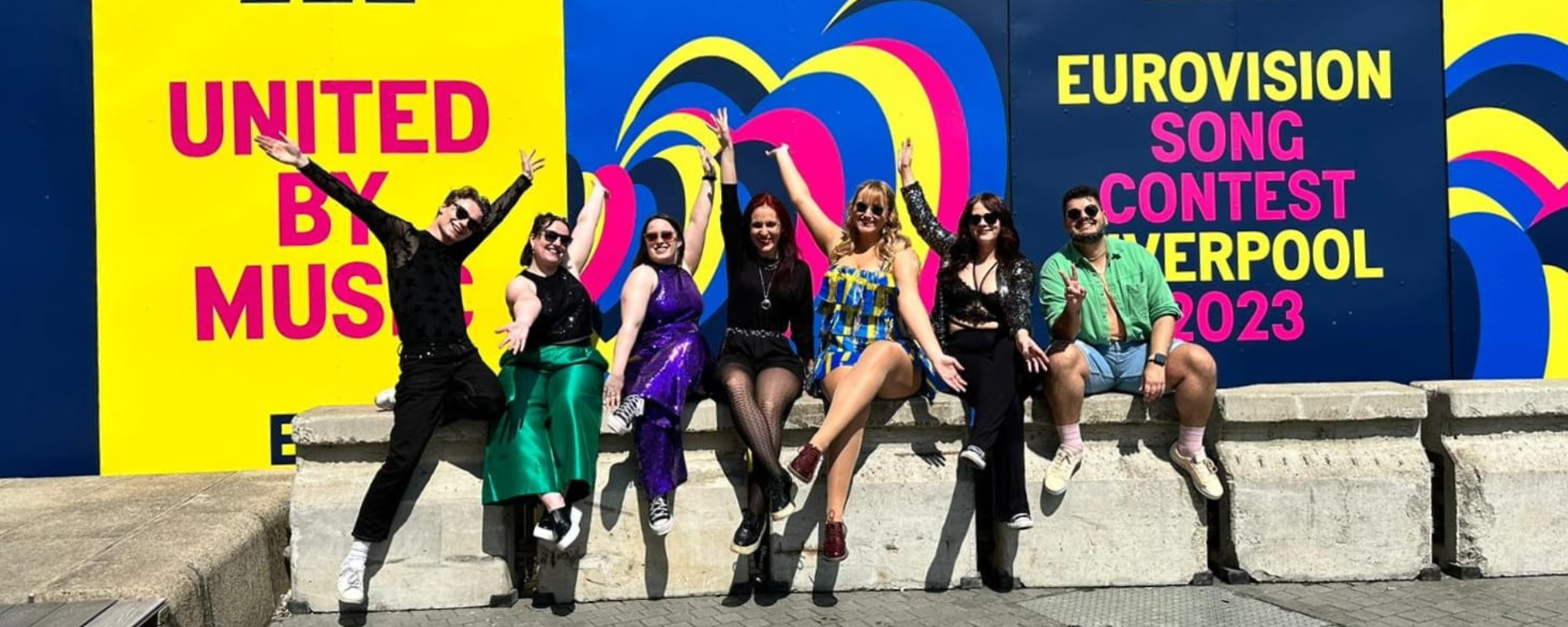 Seven lovely people sit on a wall and smile with their arms stretched out fabulously