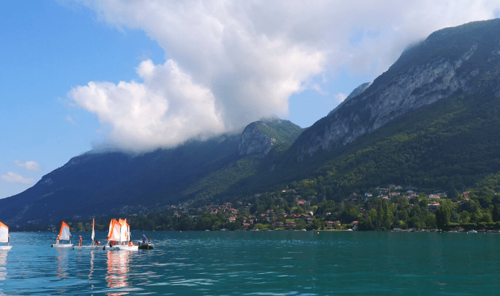 Small boats with sails sit on a bright blue lake with mountains and clouds in the background