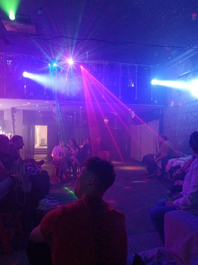 Laser party lights shine in a space, people sit on chair in a circle around the edge