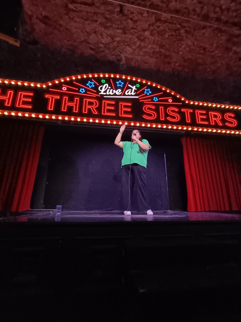 Sabina stands on a stage holding a mic as she makes us laugh