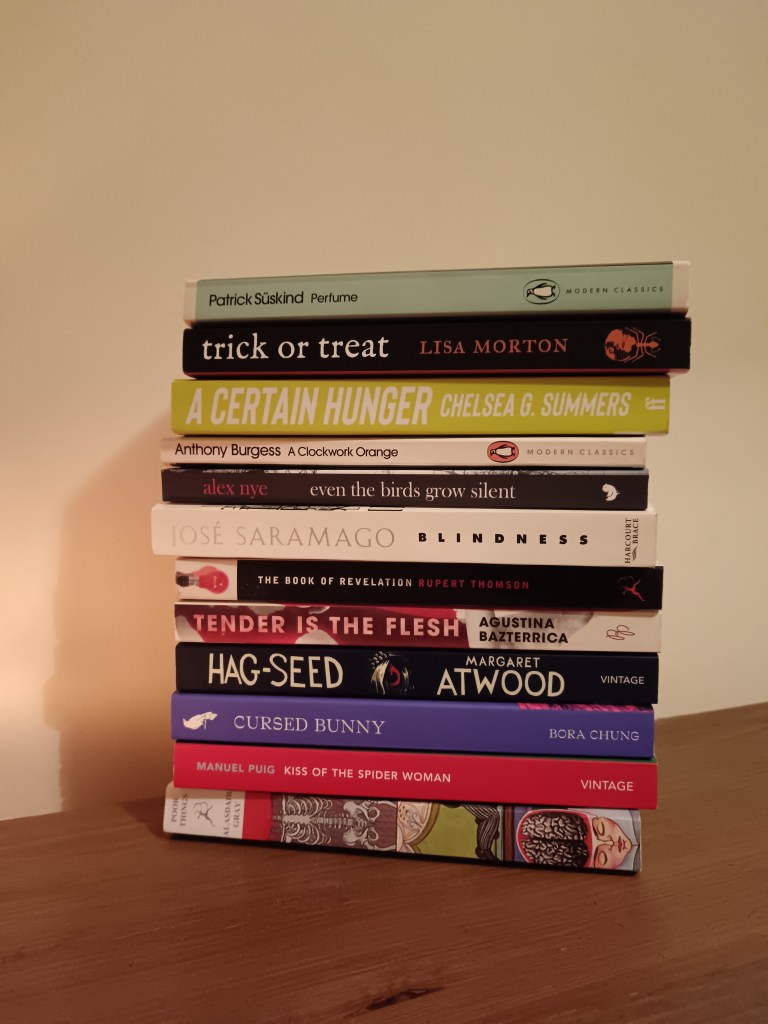 A stack of books with Perfume, Trick or Treat, A Certain Hunger, A Clockwork Orange, Even the birds grow silent, Blindness, The Book of Revelation, Tender is the flesh, Hag-seed, Cursed Bunny, Kiss of the Spider Woman and Poor Things