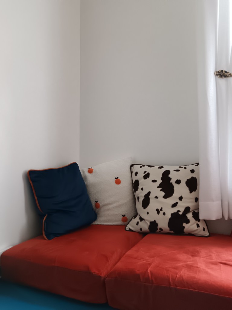 Three cushions; one blue and orange, one which with orange fruits and a cow print one