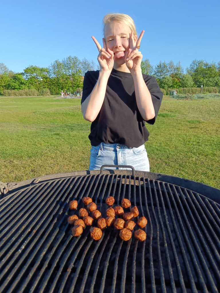 Matilda makes a peace sign standing behind meatballs on a barbecue