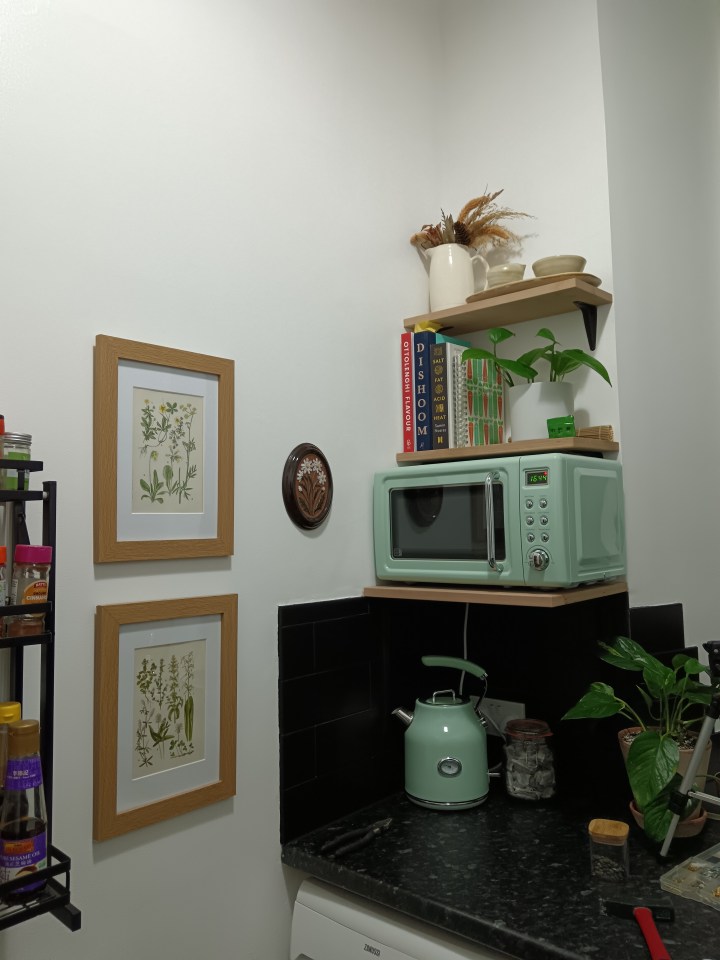 A corner of my kitchen with a sage green microwave on a wooden shelf and some plants and cookbooks. I have flower prints on the wall.