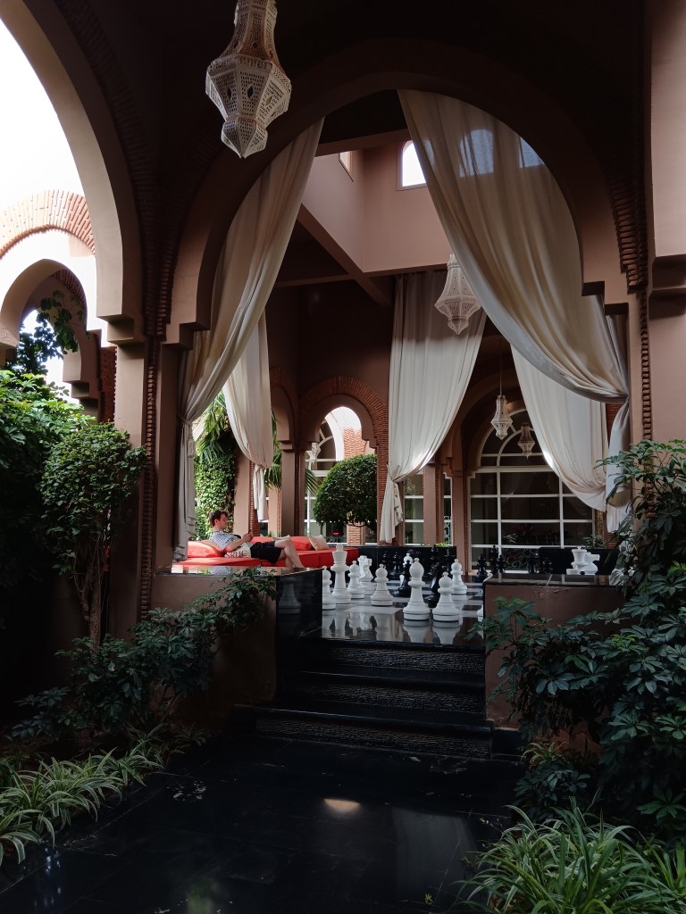 Kit sits on a sofa surrounded by fabric drapes and oversized chess pieces