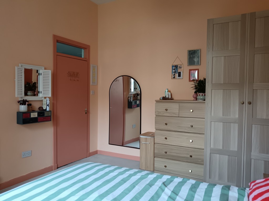 On on side of the bedroom you can see my vanity with a shuttered mirror, the door with gold hooks, a rounded black trimmed mirror, my laundry basket, oak coloured drawers and a wardrobe