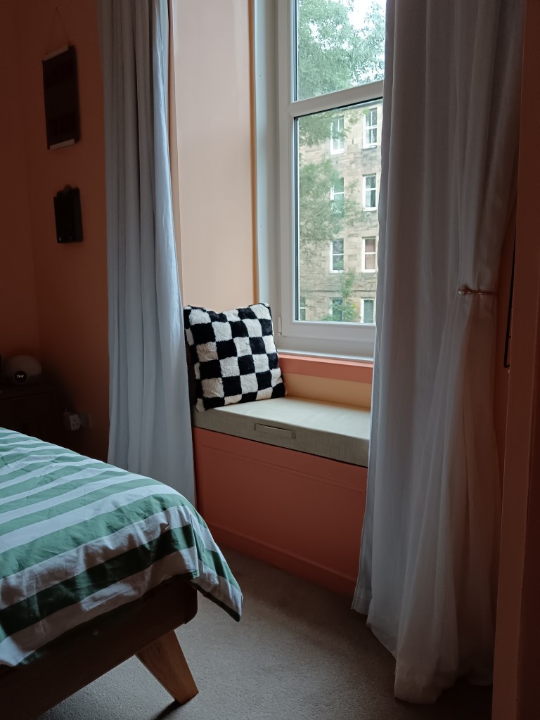 White curtains hang either side of the window and a checkered cushion sits on top of the window seat