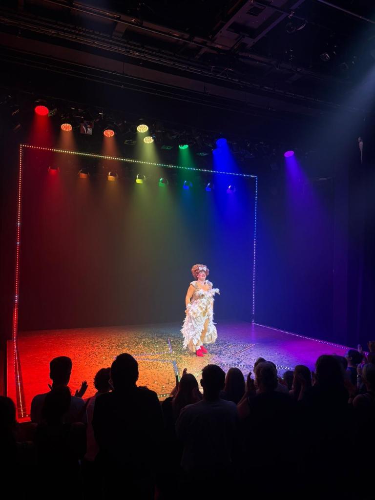 A panto dame stands in rainbow lights