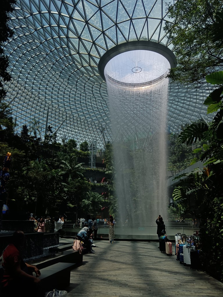 A water fountain falls from a tall ceiling in the middle of a greenhouse