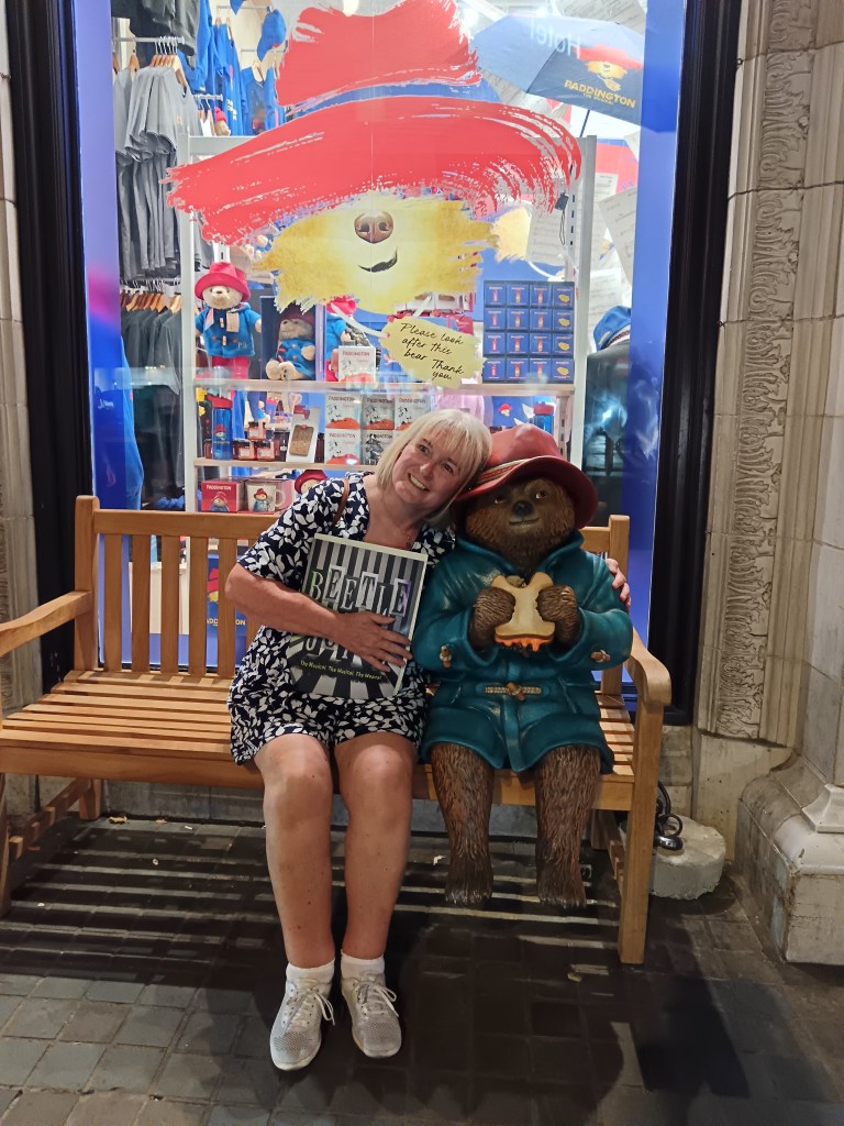 My mum smiles sitting on a bench next to a wee statue of Paddington bear