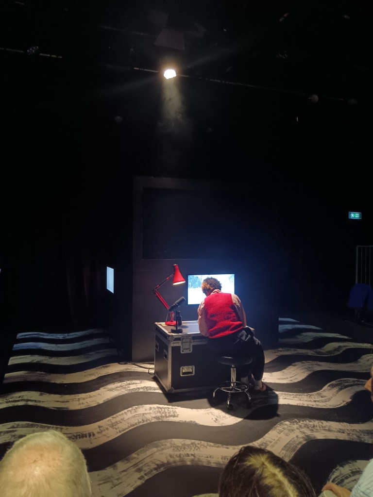 A performer sits on a stool with her back to us writing