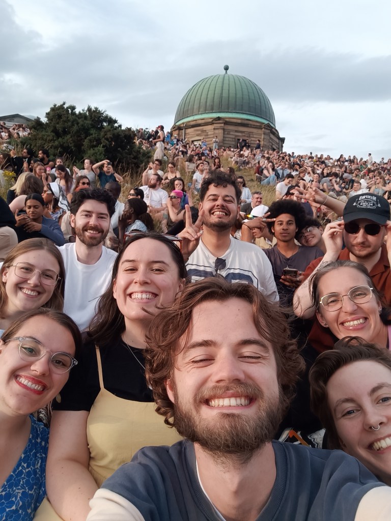 A gang of pals sit on a hill in front of the observatory
