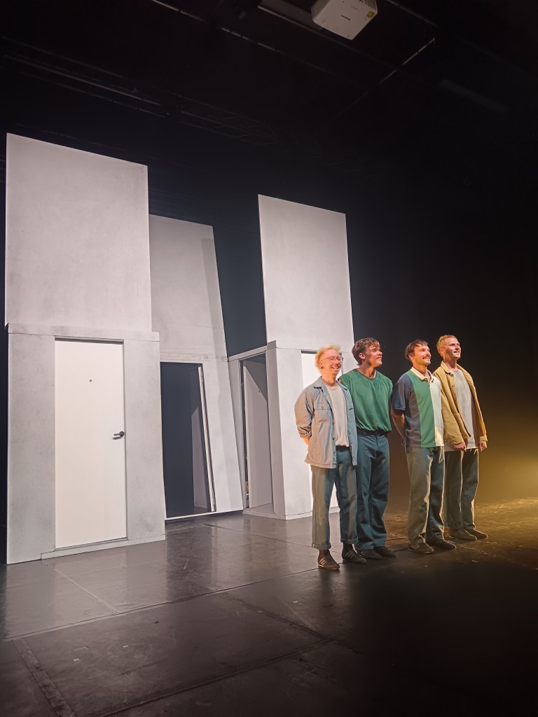Four performers smile in front of a set of grey walls and white doors