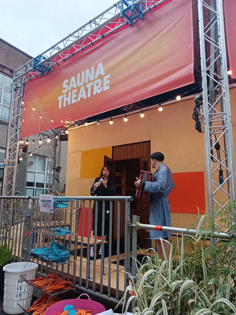 Two musicians play instruments outside the wooden Sauna Theatre building